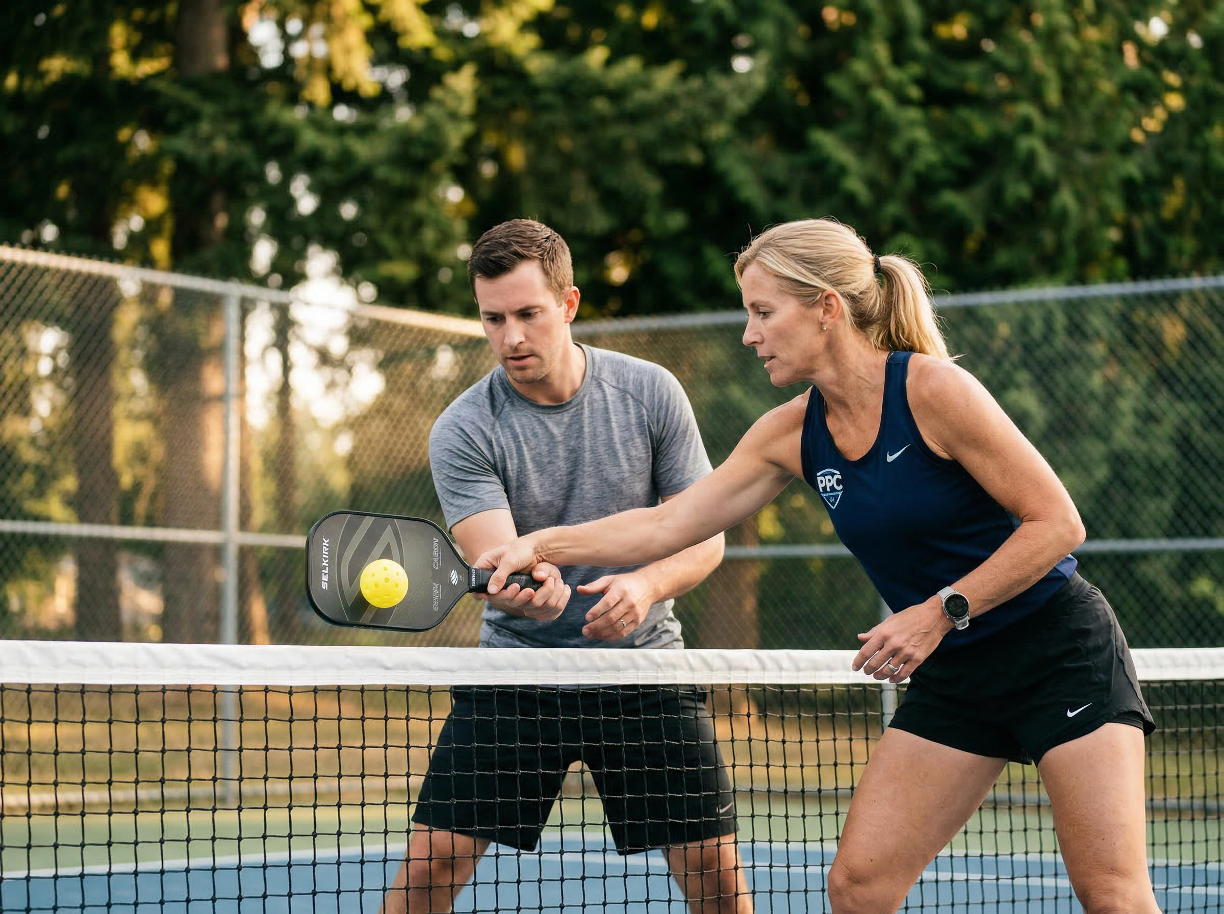 Coach demonstrating dinking technique during a private lesson on an outdoor court with Pacific Northwest trees in background